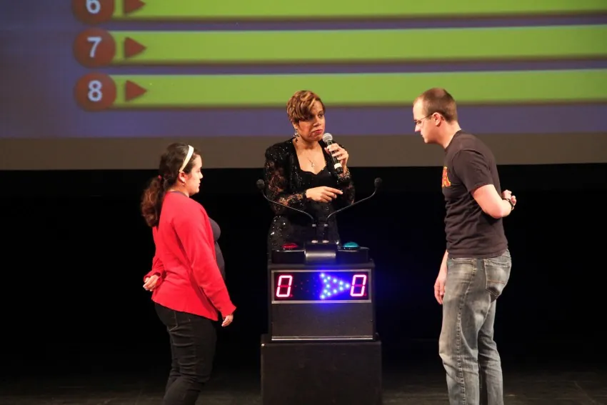 Game show host with a microphone between two contestants at the buzzer podium on stage, the answer board behind them showing slots 6, 7 and 8